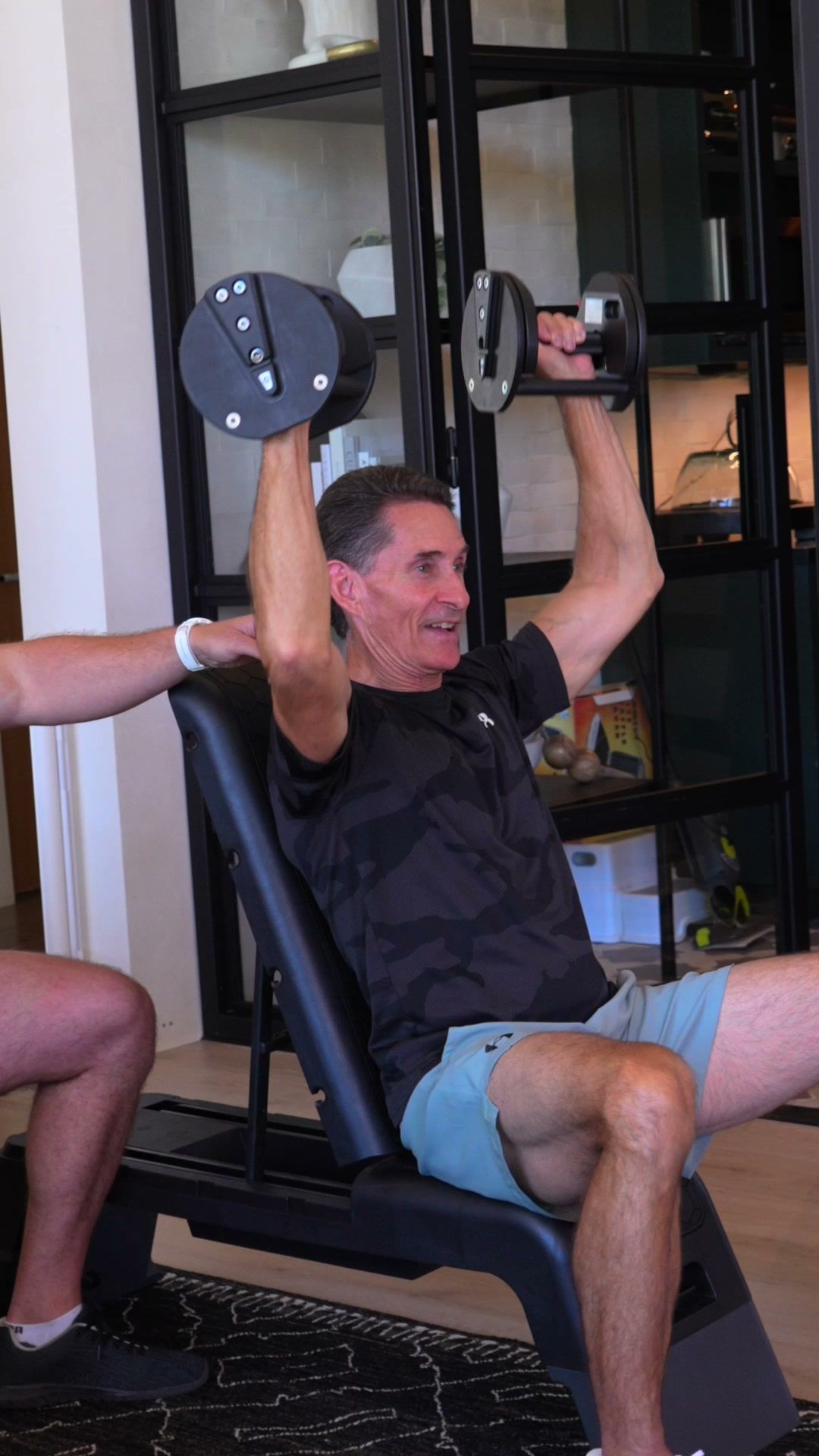 Client pressing dumbbells overhead on a bench at home
